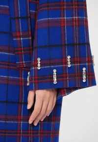 Casaco xadrez azul com detalhes em vermelho, apresentando uma textura suave e punhos embelezados com detalhes semelhantes a cristais transparentes.