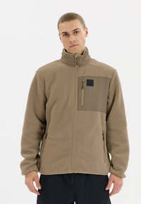 Young man with tied-back hair wearing a light brown zip-up fleece jacket with a high collar and zippered pockets, standing against a white background.