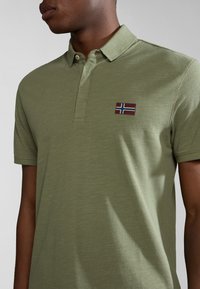 Polo in verde oliva realizzato in tessuto testurizzato, con una patta a tre bottoni e una piccola etichetta a bandiera con colori blu, rosso e bianco sul petto.