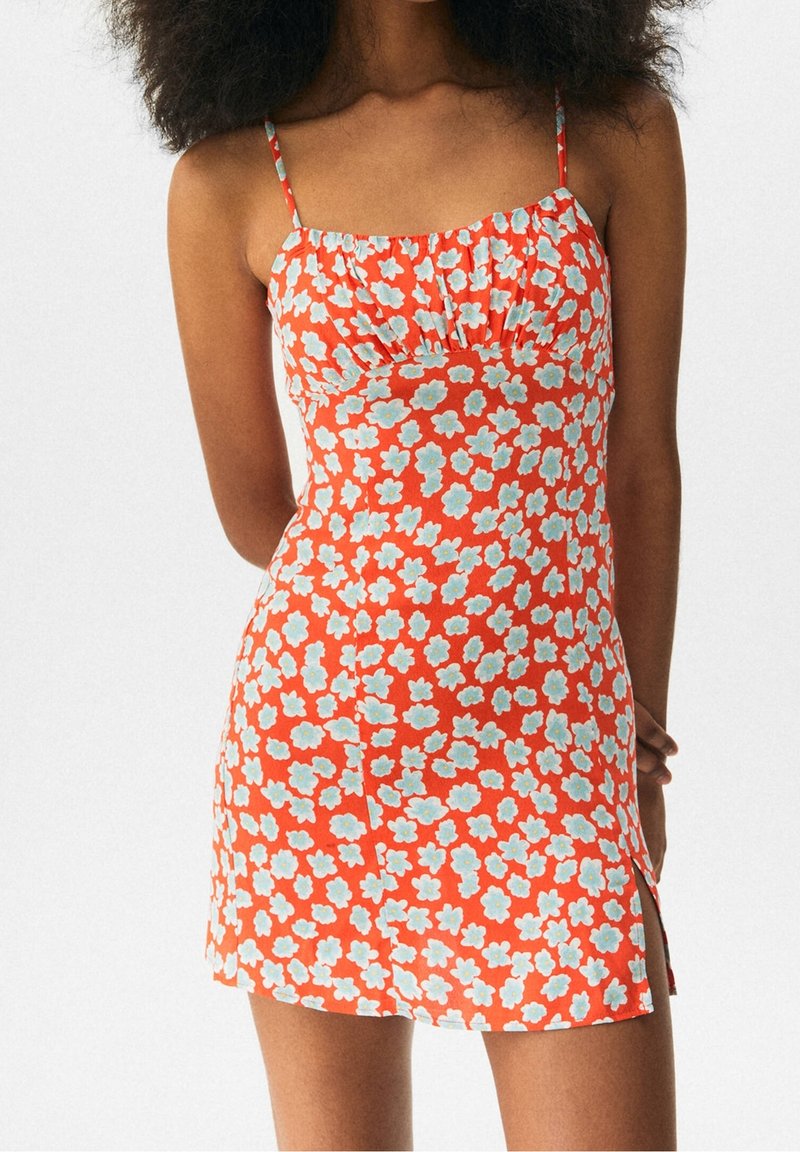 Femme portant une mini-robe orange sans manches avec un motif floral bleu et blanc et de fines bretelles, debout devant un fond blanc.