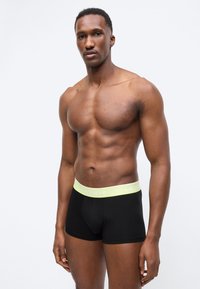 Γυμνασμένος άνδρας με σκούρο δέρμα που φοράει μαύρα εσώρουχα Lacoste boxer με κίτρινη μέση, στέκεται μπροστά σε απλό ανοιχτό φόντο.