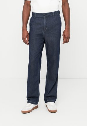 Σκούρο μπλε τζιν παντελόνι με ίσια εφαρμογή, με κούμπωμα με κουμπιά στο εμπρός μέρος, θηλιές για ζώνη και αντίθετη λευκή ραφή. Φοράει με λευκά αθλητικά παπούτσια.