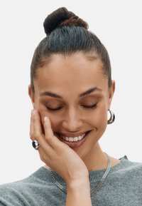 Sivý pletený sveter, vrstvený náhrdelník zo striebornej reťaze, strieborné kruhové náušnice a hladký strieborný prsteň. Vlasy upravené do drdolu. Teplý tón pleti.