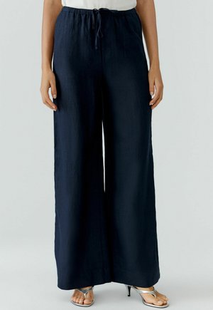 Vrouw draagt loszittende marineblauwe wijde broek met elastische tailleband en trekkoord, gecombineerd met zilveren sandaaltjes met hak.