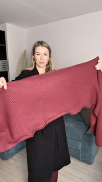 Burgundi spalvos megztas šalis, laikomas aukštyn, su tekstūruota paviršiumi ir laisvu dizainu, pasklidęs ant abiejų rankų. Fone matoma mėlyna sofa.