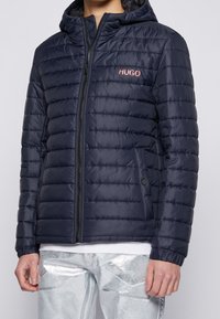 Marineblå pufferjakke med quiltet design, forreste lynlås og hætte. Har subtilt "HUGO"-logo i rød, elastiske manchetter og sidelommer.