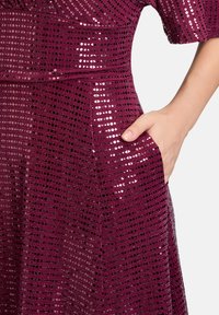 Bordowa sukienka z cekinową teksturą, krótkimi rękawami i boczną kieszenią. Posiada geometryczny wzór i dopasowany krój w talii.