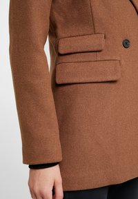 Abrigo de mezcla de lana marrón con cierre de un solo botón, diseño estructurado, tejido texturizado y dos bolsillos parche horizontales.