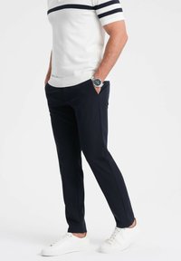 Witte polo met korte mouwen en marineblauwe strepen, gecombineerd met marineblauwe broek en witte sneakers. Horloge zichtbaar aan de linkerhand.