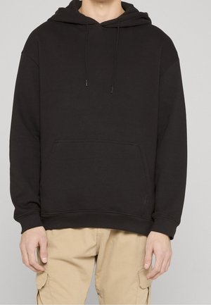 Person trägt einen schlichten schwarzen Kapuzenpullover mit Fronttasche und beige Cargohosen, steht vor neutralem Hintergrund.