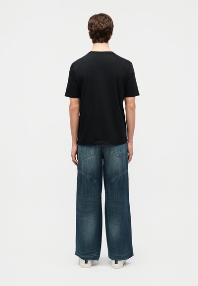 Άτομο που στέκεται με την πλάτη γυρισμένη φορώντας μια μονόχρωμη μαύρη κοντομάνικη μπλούζα, τζιν παντελόνι με φαρδιά μπατζάκια και λευκά αθλητικά παπούτσια σε λευκό φόντο.