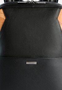 Zaino in pelle nera con superficie strutturata, dettagli in pelle color cuoio a contrasto e una placca con logo argentata sul pannello frontale. Chiusura con cerniera nella parte superiore.