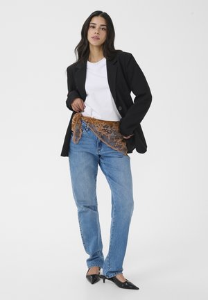 Ženska oblečena v črni blazer, belo majico, modre kavbojke s rjavo čipkasto rutko, vezano okoli pasu, in črne koničaste pete, stoji pred belim ozadjem.