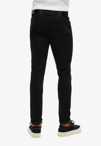 Jeans de denim negro con un corte ajustado, costuras detalladas y dos bolsillos traseros. Combinados con zapatos negros y puños remangados, mostrando calcetines naranjas.
