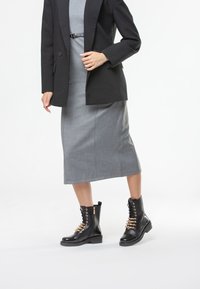 Stivaletti neri in pelle con dettagli decorativi in oro e lacci sul davanti, abbinati a un vestito midi grigio e un blazer nero.
