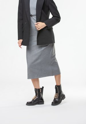 Stivaletti neri in pelle con dettagli decorativi in oro e lacci sul davanti, abbinati a un vestito midi grigio e un blazer nero.