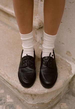 Czarne skórzane buty z zaokrąglonym noskiem, sznurowanym wzorem i teksturowaną powierzchnią, połączone z białymi, prążkowanymi skarpetkami, leżące na kamiennym stopniu.