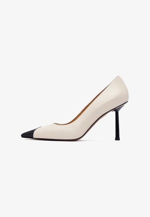 Beige leather pump with a pointed toe, black cap toe, and a slim black stiletto heel. Smooth texture and minimalist design.