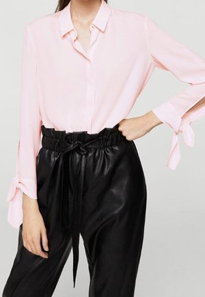 Vrouw die een lichtroze blouse met lange mouwen draagt met strikmanchetten en zwarte high-waisted paperbag-broeken met een voorstrik.