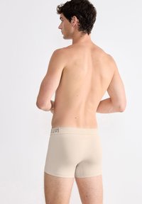 Boxers en coton beige avec une taille douce arborant un logo noir. Le modèle montre le dos, mettant en avant la coupe ajustée et la texture sans coutures.
