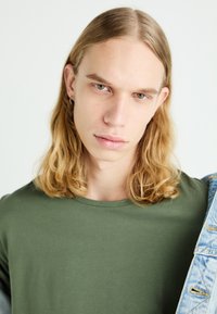 Zielona koszulka z okrągłym dekoltem wykonana z miękkiego materiału, w połączeniu z dżinsową kurtką. Modelka ma długie, falujące blond włosy i patrzy bezpośrednio w kamerę.