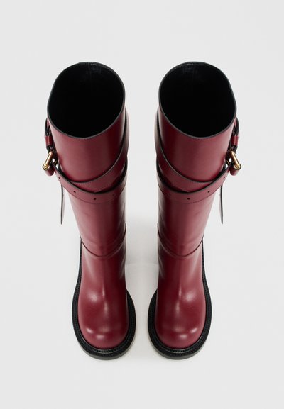 Burgundy knee-high boots made of smooth leather, featuring two black straps and a gold-tone buckle detail, with a textured black sole.