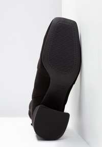 Bota de tacón alto de ante negro con punta redonda y suela de goma texturizada. Presenta costuras curvadas a lo largo de la parte superior con una base de tacón plano.