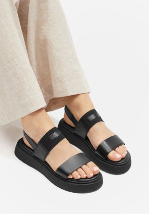 Schwarze Sandalen mit breiten Riemen und einer klobigen Sohle. Das Obermaterial ist glatt mit matter Oberfläche. Enthält elastische Akzente an der Ferse.