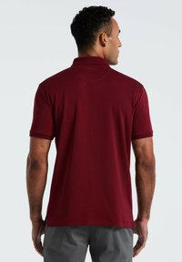 Man met kort krullend haar, gekleed in een effen bordeauxrode poloshirt en grijze broek, met de rug naar de camera toe tegen een lichtgrijze achtergrond.