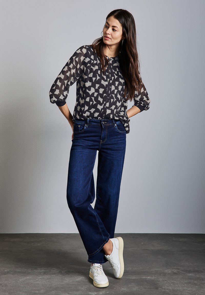 Průhledná blůza s černobílým vzorem a zapínáním na knoflíky, spárovaná s tmavě modrými volnými džínami a bílými teniskami. Hladké textury; ležérní střih.