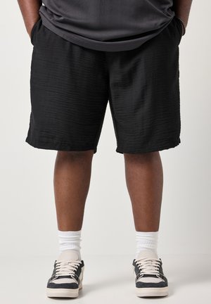 Person iført teksturerede sorte shorts med hænderne i lommerne, hvide ankelsokker og sorte og hvide sneakers, stående på en ensfarvet baggrund.