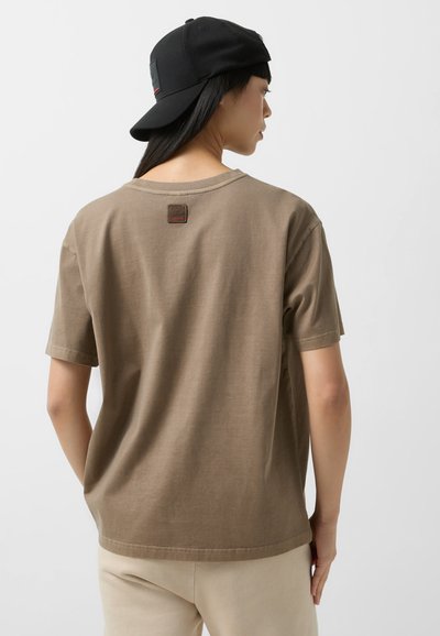 Brown cotton t-shirt with a round neck, short sleeves, and a square label on the back. Worn with a black cap and light beige pants.