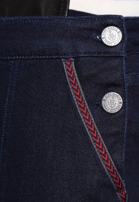 Donkerblauwe spijkerbroek heeft een rits met twee zilveren knopen, een rood chevron stiksel detail en een grijze tailleband trim.