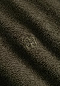 Gros plan d’un tissu tricoté vert olive foncé avec un petit logo brodé discret en fil assorti au centre.
