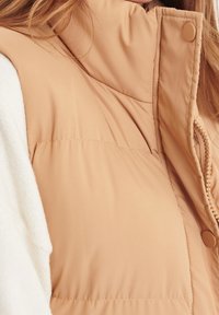 Světle hnědá péřová vesta s vysokým límečkem, zapínáním na zip a detaily na patentky. Hladký povrch s prošitými panely.