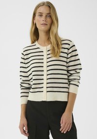 Femme aux longs cheveux blonds portant un cardigan crème avec des rayures horizontales noires et un pantalon noir, se tenant devant un fond uni.