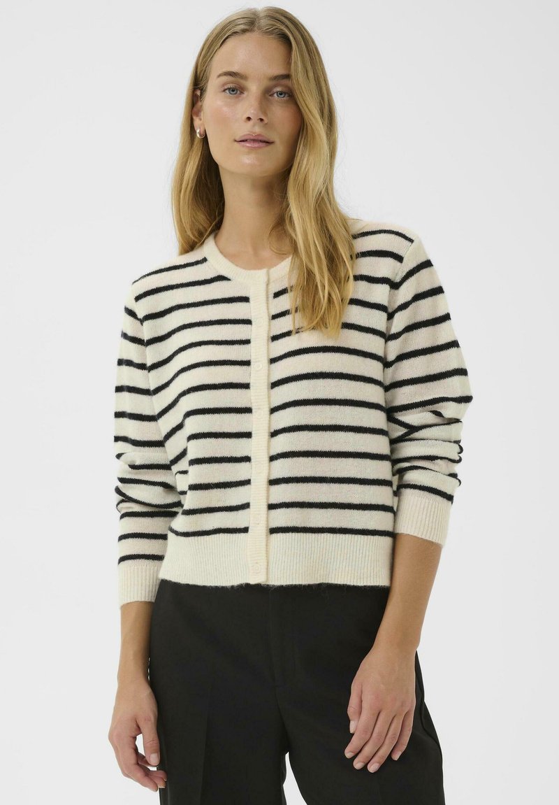 Femme aux longs cheveux blonds portant un cardigan crème avec des rayures horizontales noires et un pantalon noir, se tenant devant un fond uni.
