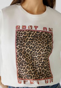 Bílé tričko s krátkým rukávem, které má na sobě grafiku se vzorem leopardího potisku v obdélníkovém rámu a tučným písmem nahoře a dole.
