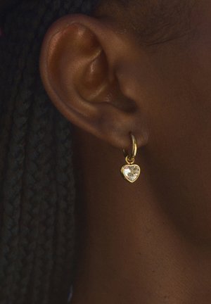 Gold heart-shaped earring with a clear gemstone, suspended from a small hoop, worn on a dark-skinned ear, highlighting detailed texture.