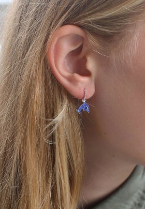 Zilveren hoopoorbel met een kleine, blauwe dolfijncharme met een gestructureerd oppervlak, gedragen aan het oor van een persoon met licht haar.