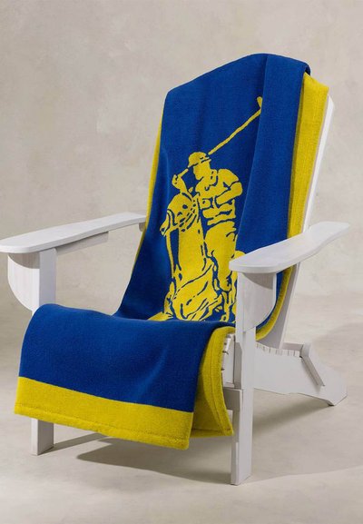 Λευκή ξύλινη καρέκλα με μια μπλε και κίτρινη πετσέτα κρεμασμένη πάνω της, με σχέδιο ενός κίτρινου παίκτη πόλο πάνω σε άλογο.
