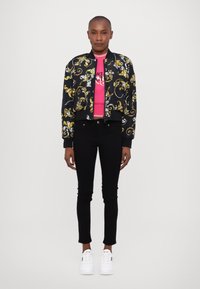 Black bomber jacket with gold and white floral patterns, pink fitted shirt, black skinny jeans, and white sneakers. Soft fabric, fitted design.