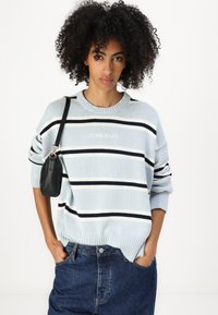Helleblaues Sweatshirt mit schwarz-weißen Streifen, gerippten Bündchen und Saum, mit besticktem Logo. Kombiniert mit dunklen Jeans und einer kleinen schwarzen Tasche.