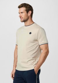 Man met lichte baard, gekleed in een beige shirt met korte mouwen, voorzien van een marineblauwe bies en een logo op de borst, en marineblauwe broek, staand met handen in de zakken.