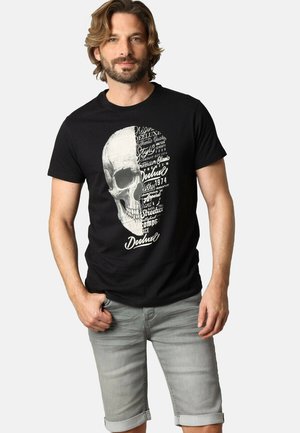 T-shirt en coton noir avec un grand graphique de crâne blanc, complété par des éléments textuels. Shorts en denim gris avec ourlets retroussés.