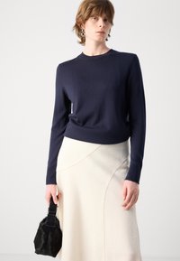 Marineblå strikket genser med rund hals, lange ermer og ribbestrikkede mansjetter, kombinert med et hvitt asymmetrisk skjørt og en liten svart håndveske.