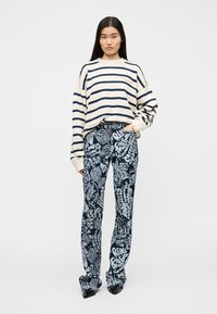 Ριγέ εκρού και ναυτικό μπλε φαρδιά ζακέτα συνδυασμένη με ναυτικά μπλε τζιν με λευκό γραφικό σχέδιο· το μοντέλο στέκεται μπροστά σε έναν απλό φόντο.