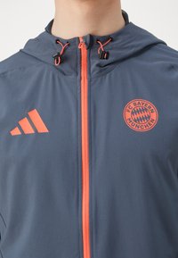 Grå treningsjakke med oransje glidelåser og aksenter. Har FC Bayern München-logo på brystet og en justerbar hette med snor.