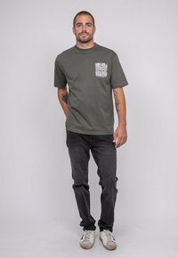 T-shirt en coton vert foncé avec un col rond et des manches courtes ; présente une poche poitrine avec du texte graphique blanc. Jeans noirs et baskets blanches.