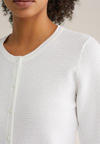 Cardigan alb cu un model texturat, cu decolteu rotund și nasturi în față. Țesătură moale cu aspect ușor.